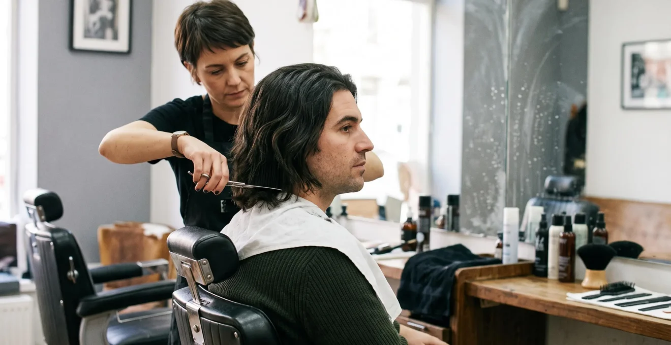 Un coiffeur structure aux ciseaux les pointes des cheveux mi-longs d'un client assis dans un salon, devant un miroir éclairé par la lumière naturelle.